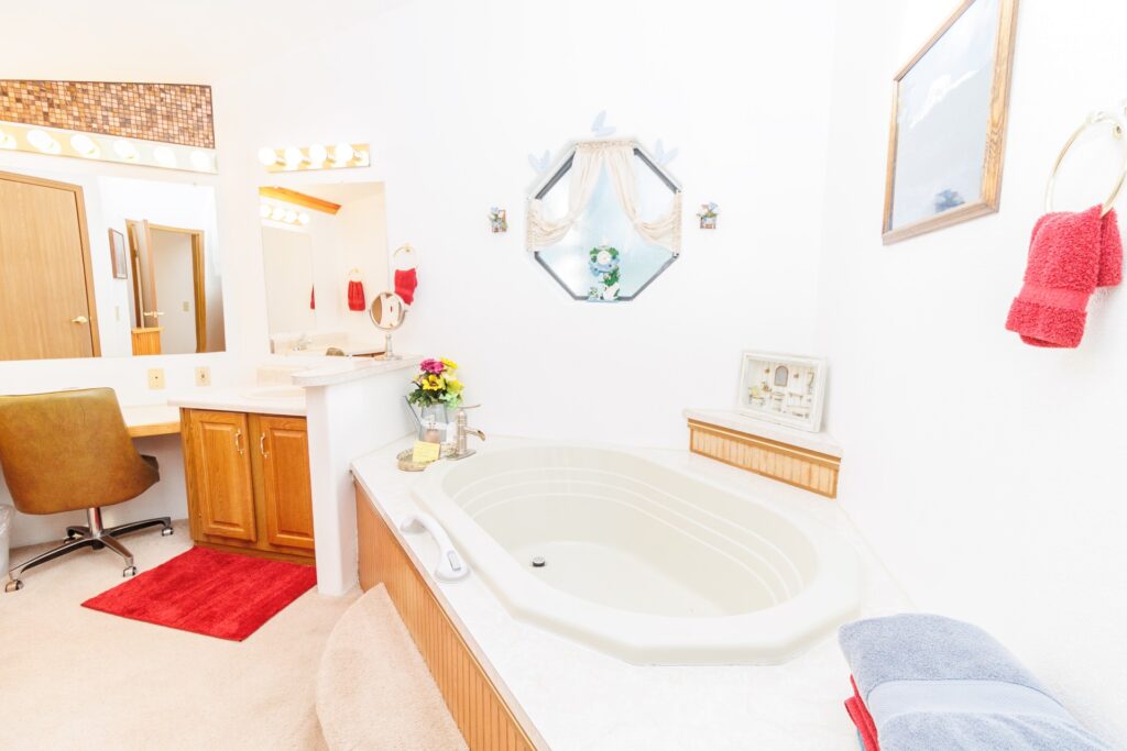 Bright bathroom featuring an oval soaking tub, a wooden vanity, and a cozy chair. Decor includes towels, flowers, and a decorative mirror.