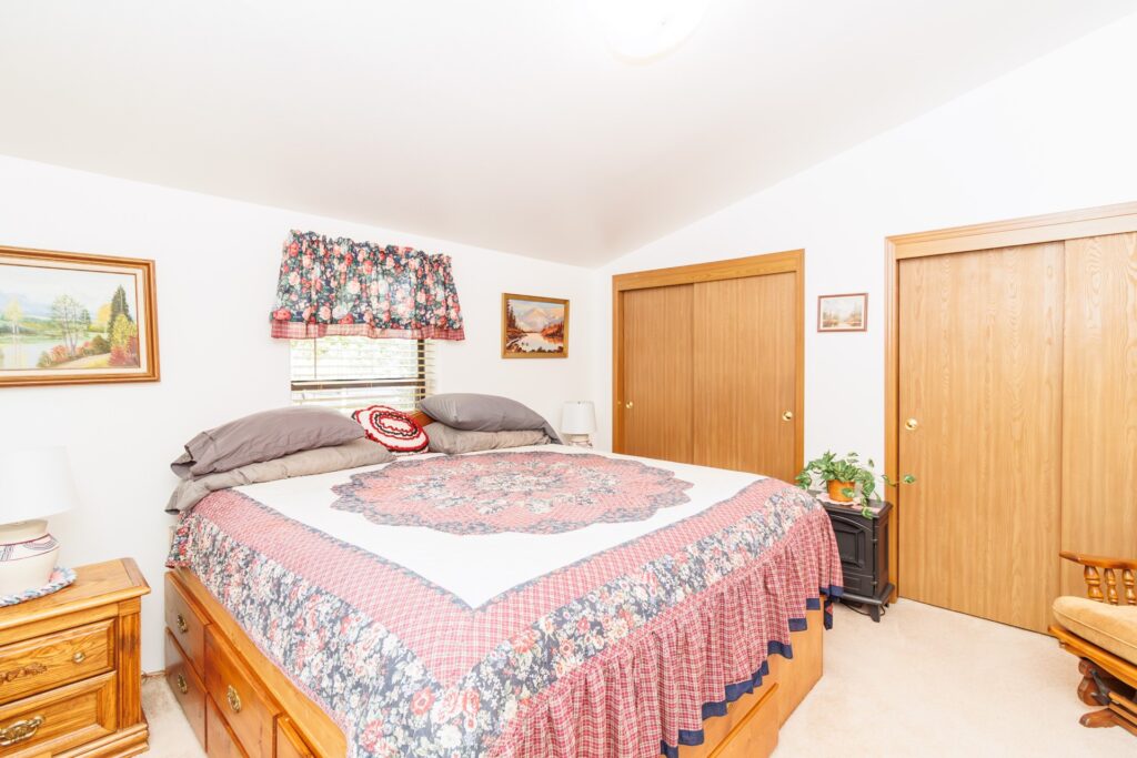 Cozy bedroom featuring a large bed with floral bedding, wooden furniture, and decorative paintings. Two closet doors and a potted plant enhance the inviting atmosphere.