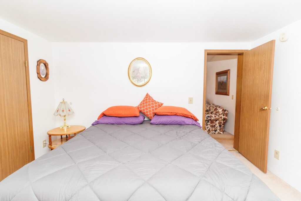 Cozy bedroom featuring a neatly made bed with colorful pillows. A lamp and decorative artwork accent the space, creating a welcoming atmosphere.