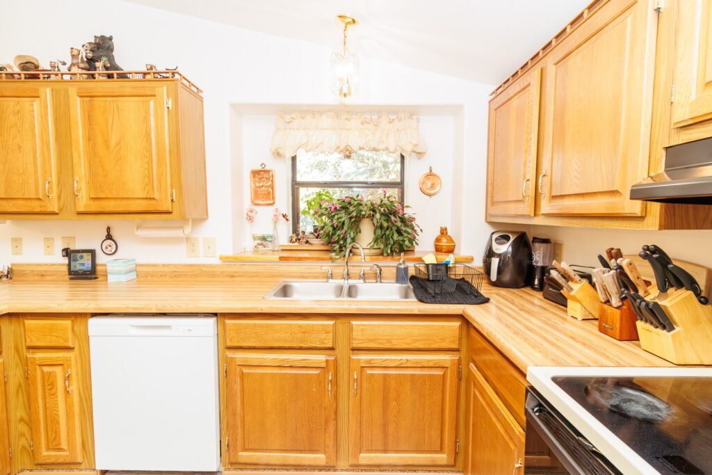 Bright kitchen featuring wooden cabinets, a double sink, and an assortment of kitchen tools. A window with plants adds natural light and charm.