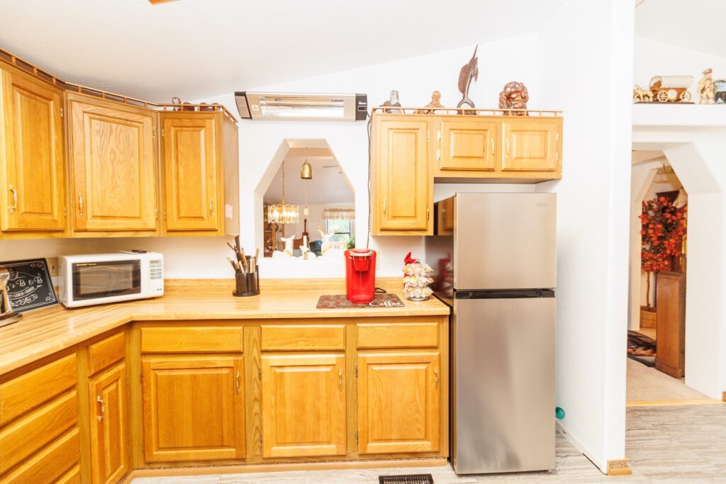 Warm-toned kitchen featuring wooden cabinets, a microwave, and a modern stainless steel refrigerator, with decorations and a view into a dining area.