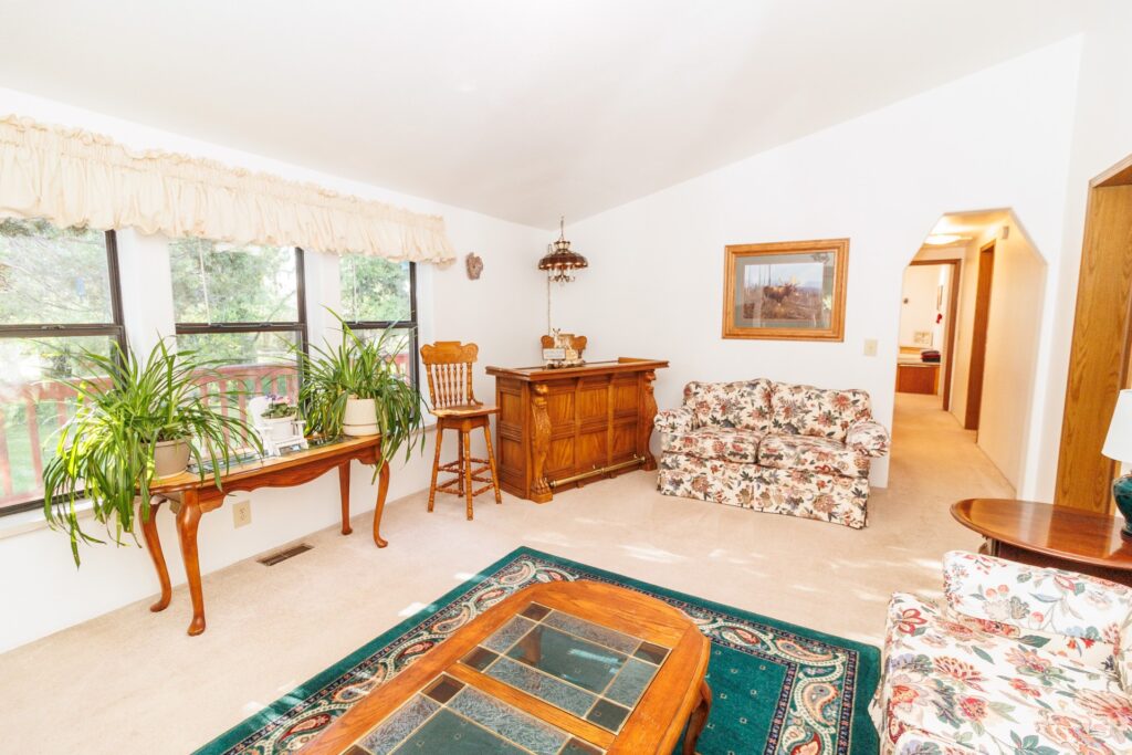 Cozy living room featuring floral couches, a wooden bar, and indoor plants by large windows, creating a bright and inviting atmosphere.