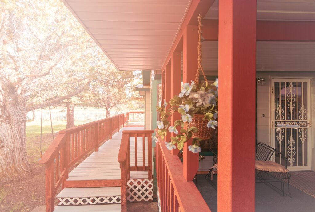 A red porch with a wooden ramp leads to a front door, adorned with a hanging flower basket; sunlight filters through surrounding trees.