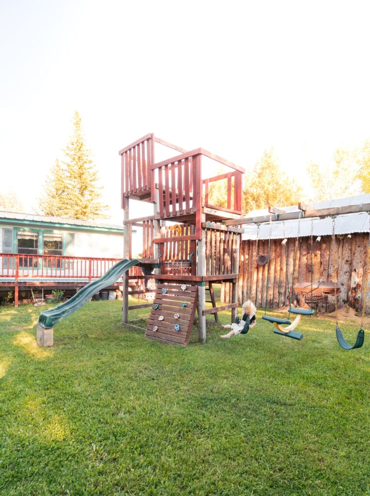 Playground with a wooden play structure featuring a slide, climbing wall, and swings, with a child playing nearby on a sunny day.