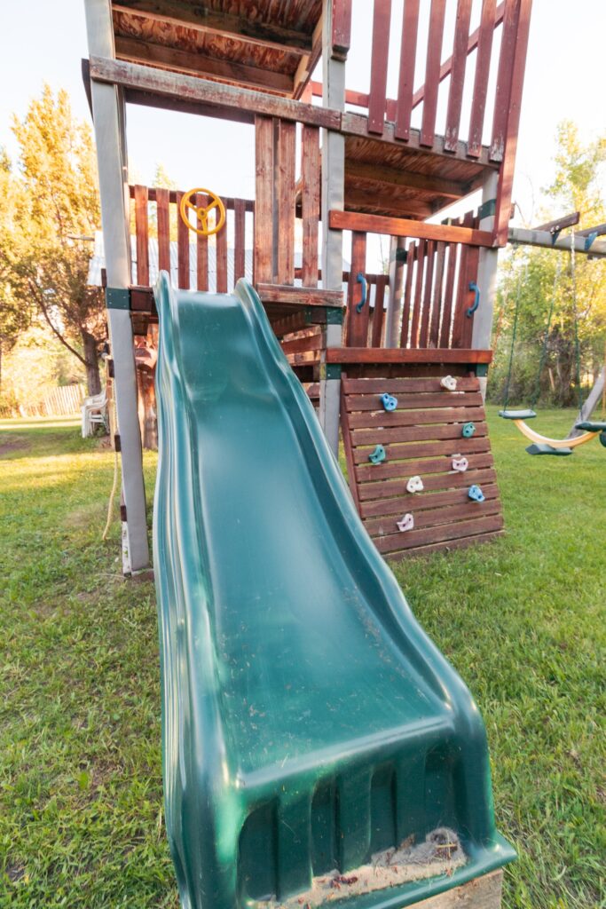 Green plastic slide attached to a wooden play structure, featuring climbing holds and a steering wheel. Set in a grassy yard, ideal for children's play.