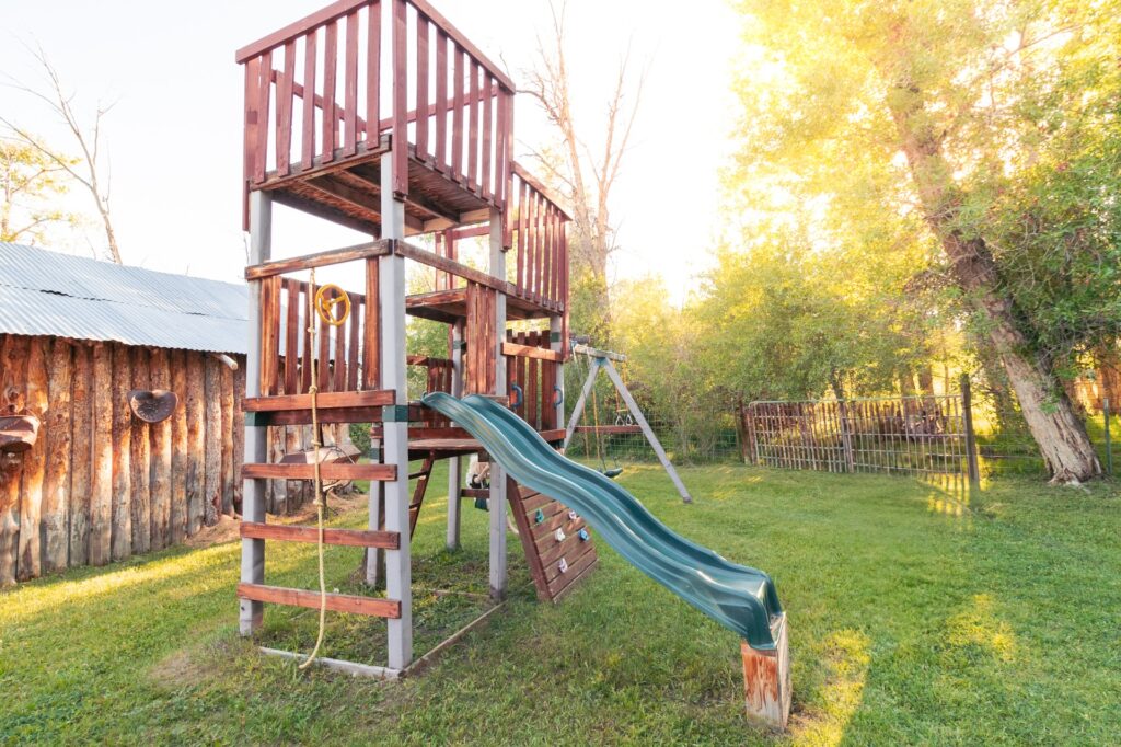 A wooden playground structure with a slide and climbing features, set in a grassy yard surrounded by trees. Ideal for outdoor children&rsquo;s play.
