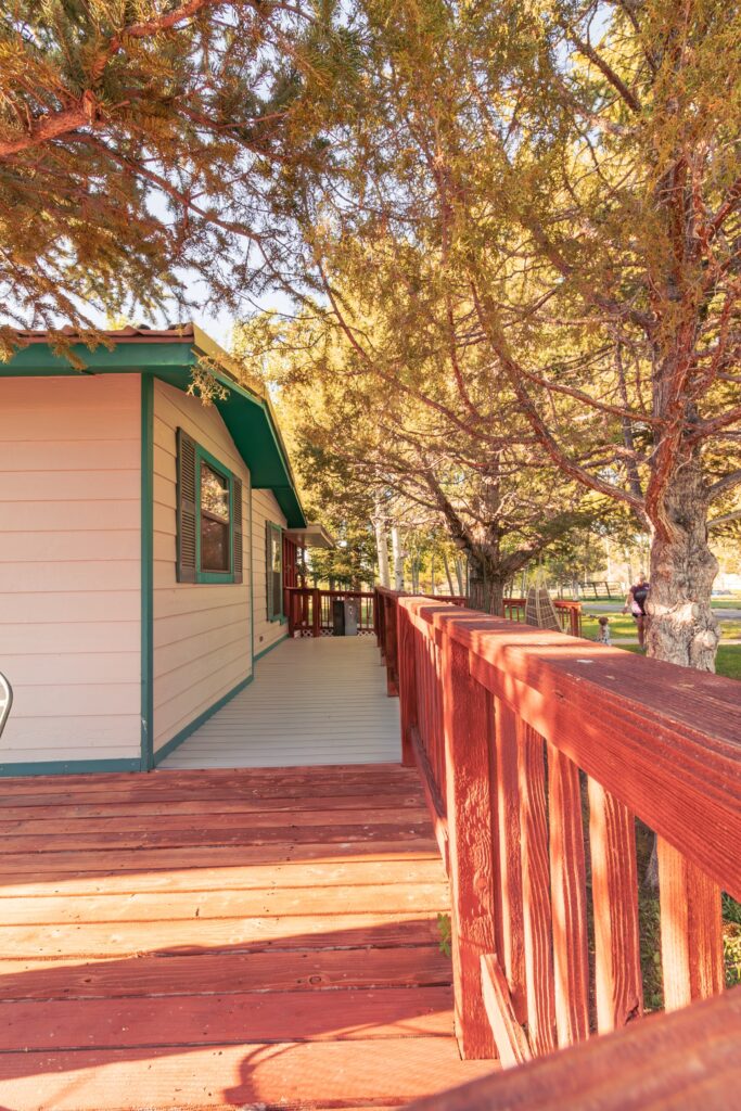 A wooden deck with a red railing leads to a cozy, light-colored cabin surrounded by lush trees, emphasizing a tranquil outdoor setting.