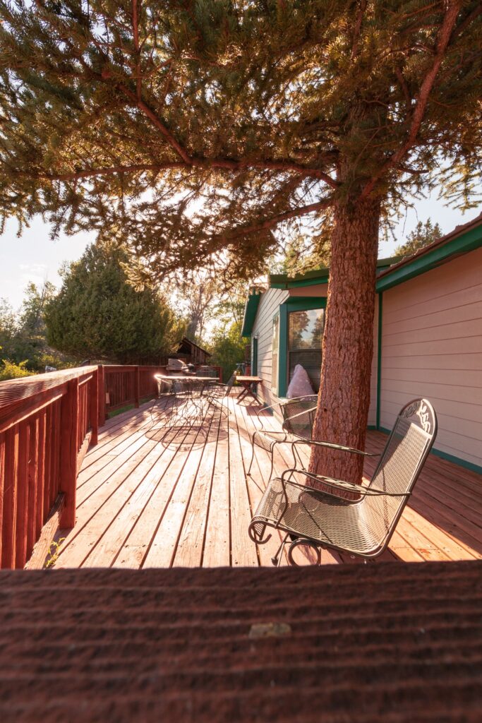 Sunlit deck surrounded by trees, featuring a wooden floor with metal chairs and a table, ideal for outdoor relaxation and gathering.