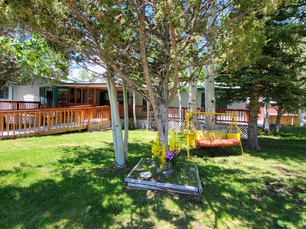 Quaint outdoor scene featuring a colorful swing beneath trees, with a wooden deck and green grass surrounding a cozy home. Ideal for relaxation.