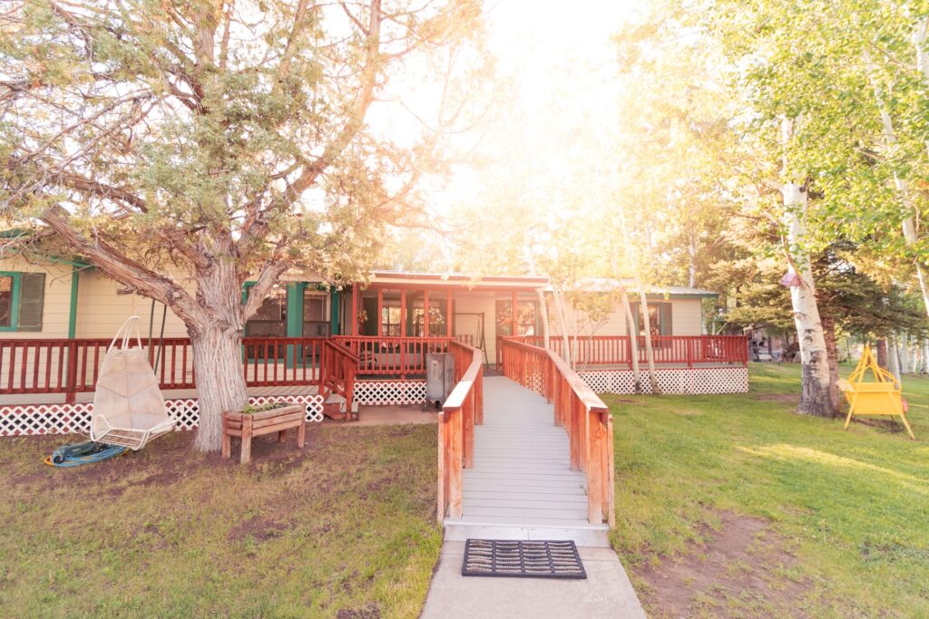 Charming single-story home with a wooden ramp leading to the entrance, shaded by trees and surrounded by lush grass, capturing a warm, inviting atmosphere.
