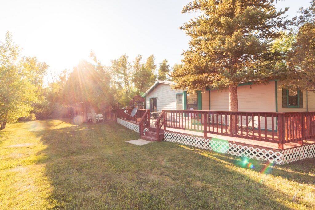 Sunlit cottage with a wooden deck and green trim, surrounded by a grassy yard and trees, creating a peaceful outdoor retreat setting.