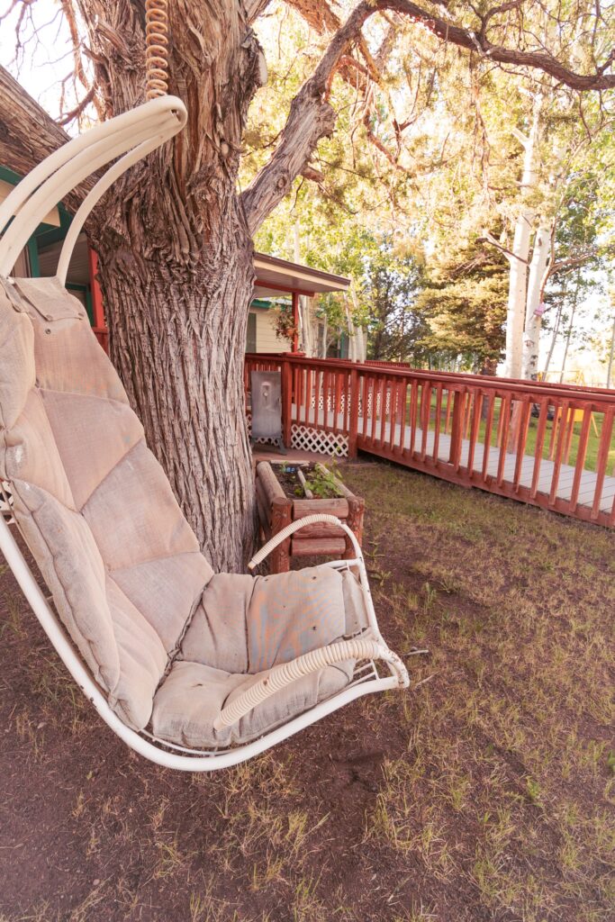 A weathered hanging chair swings from a tree, surrounded by lush green grass and a wooden deck, inviting relaxation in a serene outdoor setting.