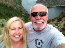 Couple smiling for a selfie at a scenic overlook, with a lake and lush greenery in the background, capturing a moment of adventure.