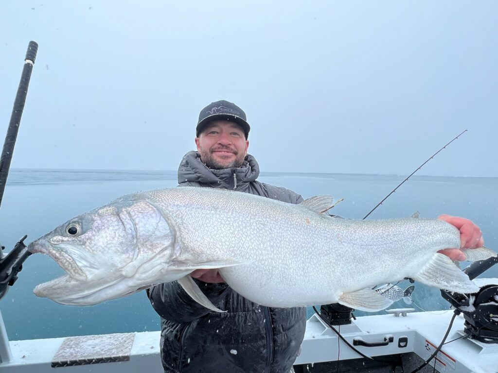 Angler proudly holds a large fish, showcasing an impressive catch on a boat in a snowy, overcast setting. The scene highlights fishing success in challenging conditions.