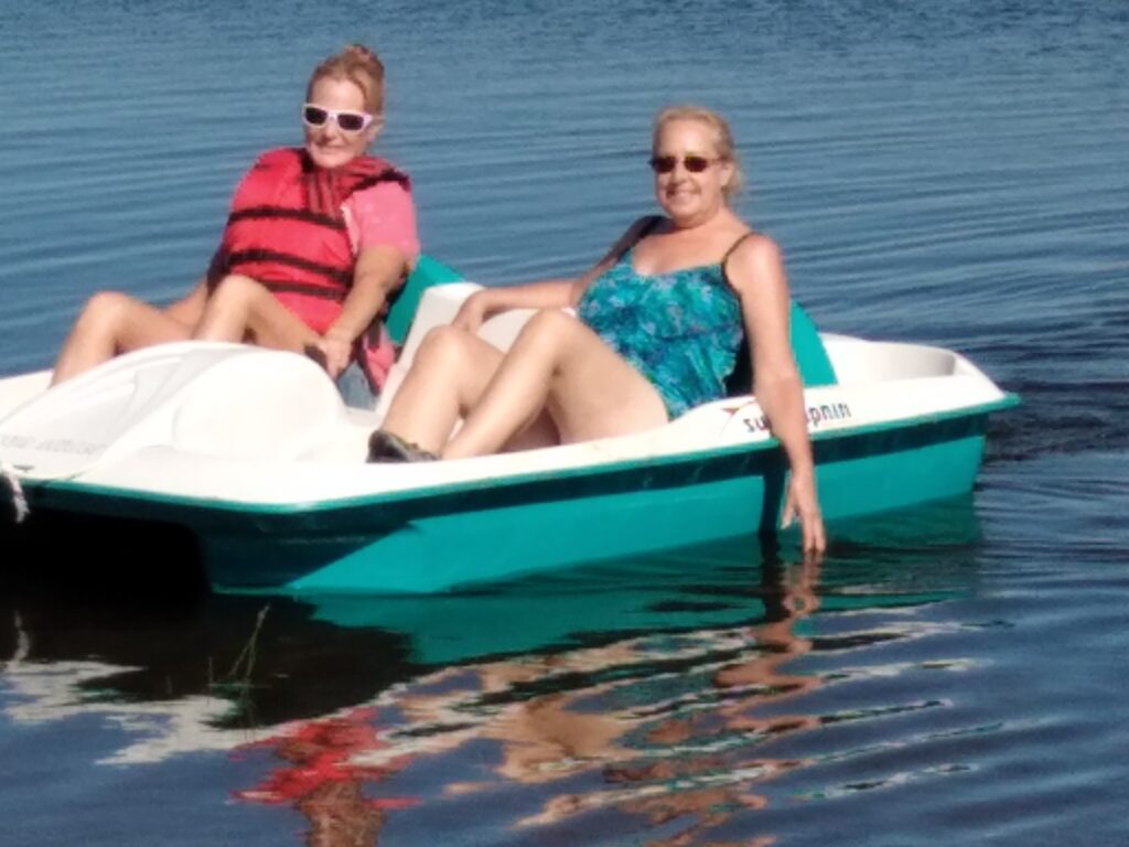 Two women relax in a pedal boat on calm water, enjoying a sunny day. One wears a life jacket and sunglasses, the other in a swimsuit.